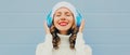 Winter portrait of happy smiling woman listening to music in headphones wearing a white hat, knitted sweater over blue Royalty Free Stock Photo