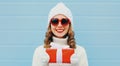 Winter portrait of happy young woman holding red gift box in her hands wearing a white knitted hat, heart shaped sunglasses Royalty Free Stock Photo