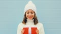 Winter portrait of happy young woman holding red gift box in her hands wearing a white knitted hat, sweater over blue Royalty Free Stock Photo