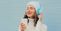 Winter portrait of smiling woman listening to music in headphones with coffee cup wearing a white hat, knitted sweater over blue Royalty Free Stock Photo