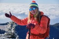 Winter portrait of woman hiker high up on a mountain, wearing insulated jacket, gloves, colorful hat and sunglasses. Sea of clouds