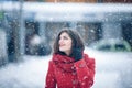 Winter portrait of young beautiful brunette woman wearing knitted snood and red coat covered in snow. Snowing winter beauty fashio