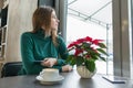 Winter portrait of young beautiful girl sitting at cafe with cup of coffee and mobile phone, snow outside the window, Christmas