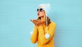 Winter portrait young woman blowing red lips sending sweet air kiss wearing yellow knitted sweater and white hat with pom pom Royalty Free Stock Photo