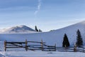 Winter rural background with small wooden alpine house, pines, fence, snow field, mountains, blue sky Royalty Free Stock Photo