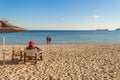 Winter sea beach and calm sea on a sunny day. A lonely elderly woman sits in a deck chair facing the sea. A loving couple stands Royalty Free Stock Photo