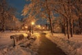 Winter tale in city park with snow covered trees, wooden benches and row of lamps along alley at the night
