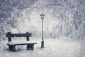 Winter time scene with a wooden bench and street lamp covered with snow under a snowfall in the park. Wonderful holiday season Royalty Free Stock Photo