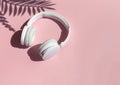 Wireless white headphones with tropical leaves shadows background on the pink close up. Listening to summer music concept.