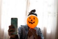 Witch holding a pumpkin balloon with her hand, covering her face and taking a photo with mobile phone. Young woman celebrating Royalty Free Stock Photo