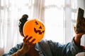 Witch holding a pumpkin balloon with her hand, covering her face and taking a photo with mobile phone. Young woman celebrating