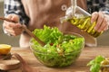 Woman adding olive oil into bowl with fresh vegetable salad on table