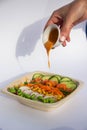 Woman adding sauce to tasty poke bowl with fresh vegetables and other ingredients on white background from side view