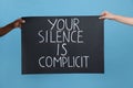 Woman and African American man holding sign with phrase Your Silence Is Complicit on light blue background, closeup. Racism