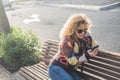 Woman alone in the street sitting on a bench of the park using her phone and smiling at it - online lifestyle concept - curly Royalty Free Stock Photo