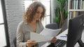 Woman analyzing documents at office desk in modern workplace with brick wall and plants, focusing on work with computer, Royalty Free Stock Photo