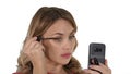 Woman applying black mascara on eyelashes looking in her phone on white background. Royalty Free Stock Photo