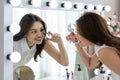 A woman is applying makeup while looking into vanity mirror with light on a table. She is using an eyelash curler and has a Royalty Free Stock Photo