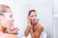 Woman applying mask moisturizing skin mask on face looking in bathroom mirror. Girl taking care of her complexion layering Royalty Free Stock Photo