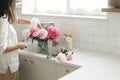 Woman arranging beautiful peonies in vase at sink with brass faucet in new modern kitchen. Female decorating flowers on background Royalty Free Stock Photo