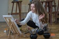 Woman artist sits on the floor among easels with paintings in an art studio and paints an oil painting Royalty Free Stock Photo