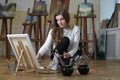 Woman artist sits on the floor among easels with paintings in an art studio and paints on canvas Royalty Free Stock Photo