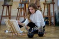 Woman artist sits on the floor among easels with paintings in an art studio and paints an oil painting Royalty Free Stock Photo