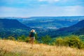 A woman as a tourist takes pictures and looks at a beautiful valley with green trees, a forest on a hill and wild grass, a blue