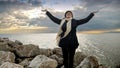 Woman with a backpack stands on a sea cliff, arms stretched out, enjoying the magnificent view of cold ocean waves. Perfect for