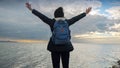 Woman with a backpack stands at a sea cliff, taking looking at the cold ocean waves with arms outstretched. This footage is ideal Royalty Free Stock Photo