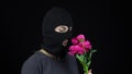 A Woman in a balaclava mask is standing with flowers. Bandit sniffs a bouquet of pink flowers on a black background.