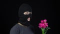 A Woman in a balaclava mask is standing with flowers. Bandit sniffs a bouquet of pink flowers on a black background.