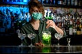 Woman bartender in medical mask mixes green drink in glass using spoon.