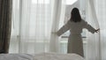 Young woman in bathrobe unveil curtains and looks from the window