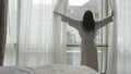 Young woman in bathrobe unveil curtains and looks from the window