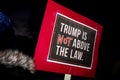 Woman in beanie cap and fur collar holds sign Trump is NOT Above the Law at night protest