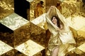 Woman beauty portrait of fashion model in golden dress, waving golden glittering fabric flying in front of cube background Royalty Free Stock Photo