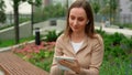 Woman in a beige lingo wearing wireless headphones uses a tablet while sitting on a bench in the park