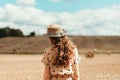 Woman in beige polka dot dress with curly hair, straw hat standing on harvested field with straw bales. Agriculture background