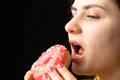 A woman bites a large red donut, a black background, a place for text. Gluttony, overeating and sugar addict.