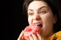 A woman bites a large red donut, a black background, a place for text. Gluttony, overeating and sugar addict.