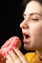 A woman bites a large red donut, a black background, a place for text. Gluttony, overeating and sugar addict.