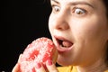 A woman bites a large red donut, a black background, a place for text. Gluttony, overeating and sugar addict.