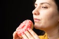 A woman bites a large red donut, a black background, a place for text. Gluttony, overeating and sugar addict.