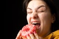 A woman bites a large red donut, a black background, a place for text. Gluttony, overeating and sugar addict.