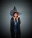 Woman with black cape and witch hat place two hands under her face on stripe metal background Royalty Free Stock Photo