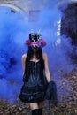 A woman in a black dress with a corset and a top hat decorated with skeleton figures poses for the camera in blue smoke