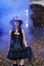 A woman in a black dress with a corset and a top hat decorated with skeleton figures poses for the camera in blue smoke