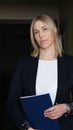 woman in a black formal blazer holding documents and looking at camera. blond confident woman business boss standing