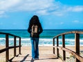 A woman with black hair taken from behind with a tourist backpack, walking towards the beach on a wooden path, arms retracted